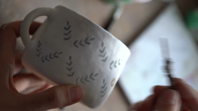 Woman paints mustard circles on white ceramic mug using brush for pottery on blurred background