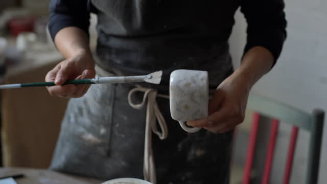 Female artisan paints white ceramic cup with brush standing in studio on blurred background