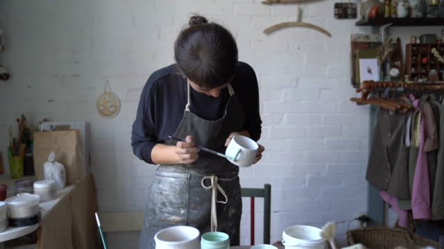 Young brunette woman in black apron paints white ceramic mug standing against brick wall in workshop