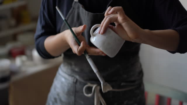 Woman in black apron enjoys new creative hobby painting white ceramic mug with brush in workshop