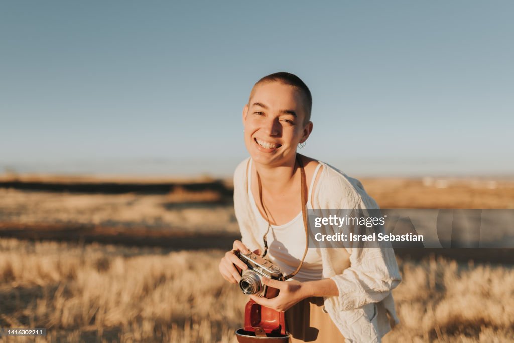 Woman with shaved hair laughing out loud and holding with a vintage camera in the field at sunset.