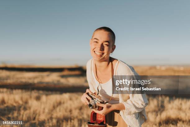 woman with shaved hair laughing out loud and holding with a vintage camera in the field at sunset. - shaved head stockfoto's en -beelden