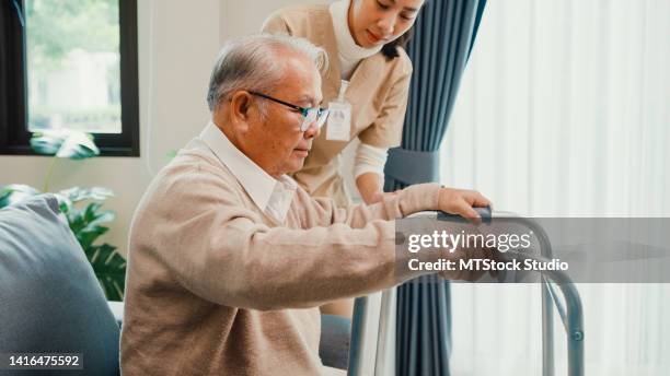 young asian female caregiver teach old male to walk use walker take care hold hand elderly patient in living room at home. - hip replacement stock pictures, royalty-free photos & images