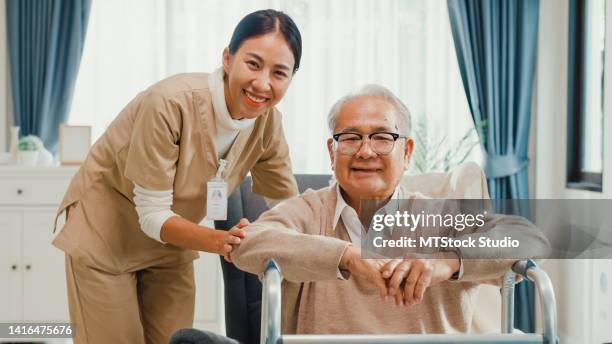 happy life asian retirement senior older male sit on wing chair with walker and female nurse healthcare giver hold hand smiling looking at camera at home. - better decisions stock pictures, royalty-free photos & images