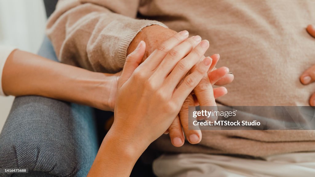 Close-up Young Asian female caregiver hold old male hand take care elderly patient on wing chair at home.