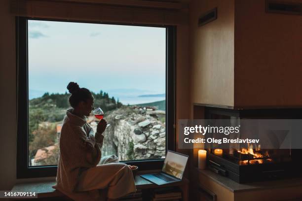 woman relaxing in evening, drinking red wine and watching movies on laptop near fireplace. - kaminabend stock-fotos und bilder