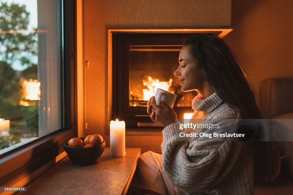 Smiling woman in woolen sweater drinking warm tea at home near fireplace