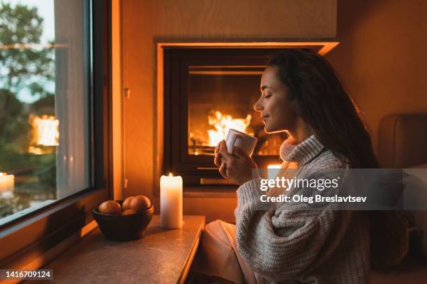 smiling woman in woolen sweater drinking warm tea at home near fireplace - hygge bildbanksfoton och bilder