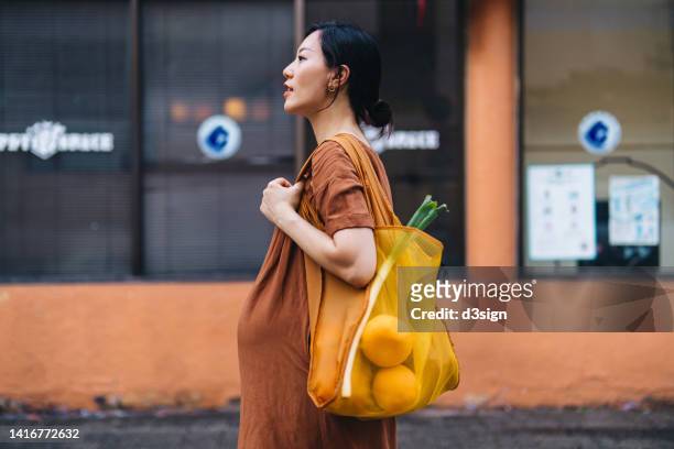 young asian pregnant woman carrying a yellow reusable shopping bag, shopping for fresh organic fruits and groceries in the city, standing against orange wall. responsible shopping, zero waste, sustainable and healthy eating lifestyle during pregnancy - fair trade stock-fotos und bilder