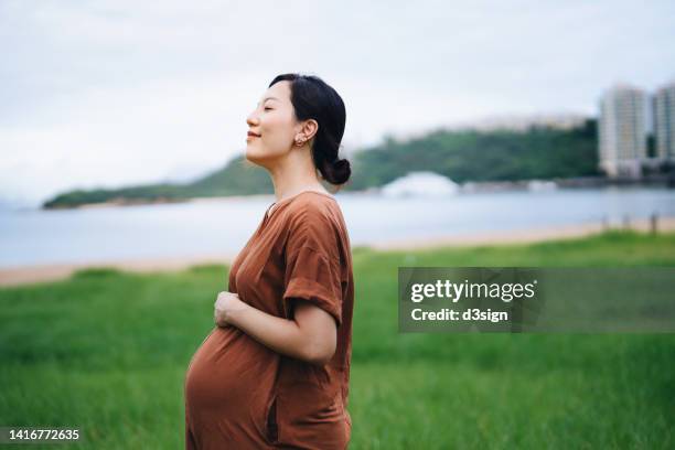 beautiful young asian pregnant woman with eyes closed standing in the meadow, gently touching her baby bump. breathing fresh air and enjoying nature with smile. mother-to-be. enjoying her pregnancy journey. love and bonding. healthy pregnancy lifestyle - breathing new life stock pictures, royalty-free photos & images