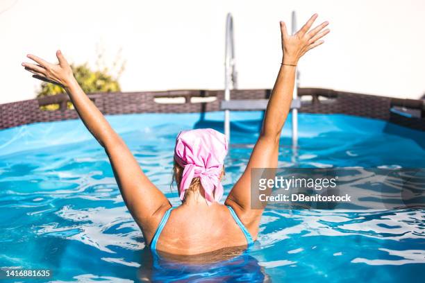 female breast cancer survivor, wearing a swimsuit in a swimming pool, wearing the pink scarf to fight breast cancer and raising her hands. concept of disease, chemotherapy and success. - best bosom fotografías e imágenes de stock