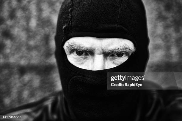 scary man frowns and stares at camera, wearing black balaclava and leather jacket - bivakmuts stockfoto's en -beelden