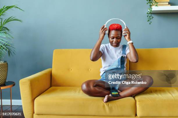 music is life. young african american woman putting on headphones, sitting on sofa in lotus pose - red telephone box stock-fotos und bilder