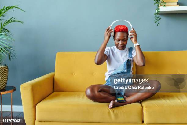 music is life. young african american woman putting on headphones, sitting on sofa in lotus pose - 21 & over stock pictures, royalty-free photos & images