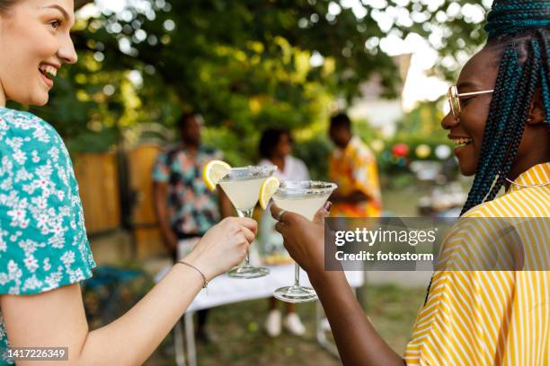 candid shot of two diverse girlfriends cheering with margarita cocktails - summer cocktails garden party drinks stockfoto's en -beelden