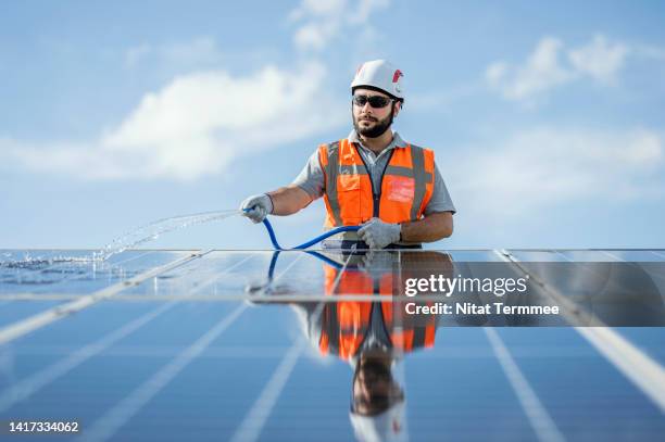 water consumption in pv panel cleaning and maintenance. low-angle view of a solar technician spraying water for clean solar panels following maintenance plan in a solar power plant. - power grid stock-fotos und bilder