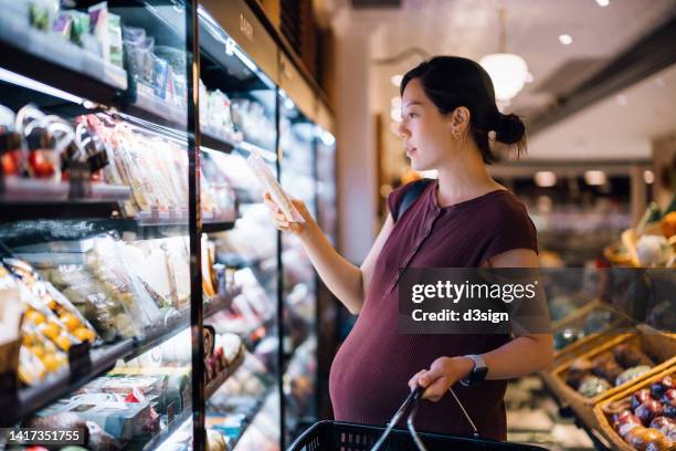 young asian pregnant woman grocery shopping in supermarket, choosing fresh packets of cheese from the diary aisle. eating well with balanced nutrition. pregnancy health and wellness. healthy eating habit and lifestyle during pregnancy - vitamins and minerals imagens e fotografias de stock