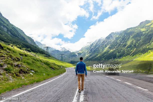 rear view of a man walking on the country road surrounded by mountains - romania stock pictures, royalty-free photos & images