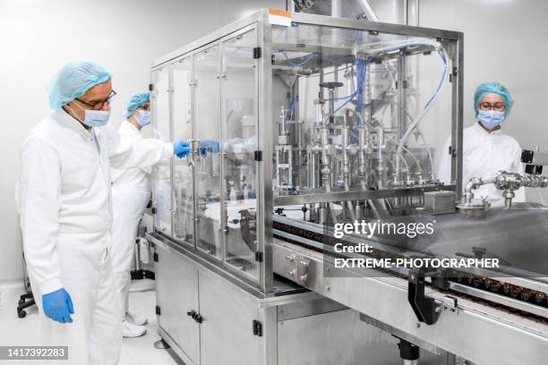 tres colegas farmacéuticos vistos mientras trabajaban en una máquina que está llenando las botellas durante el proceso de fabricación - fábrica farmacéutica fotografías e imágenes de stock