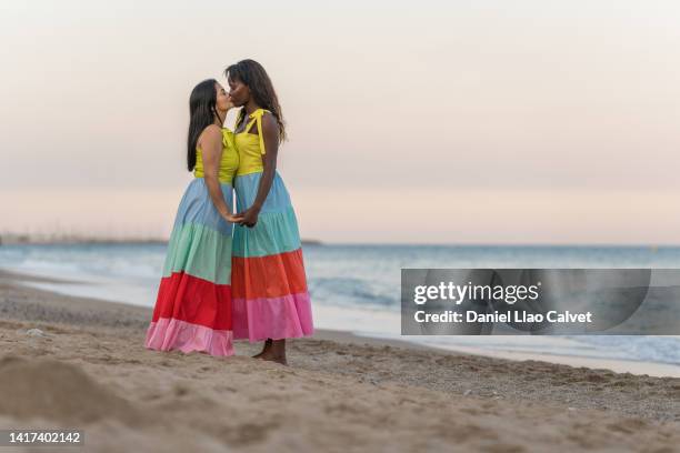 two women with colourfull dresses hold hands while kissing on a beach. - photos of lesbians kissing stock pictures, royalty-free photos & images