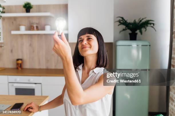 smiling woman holding illuminated light bulb in kitchen at home - gloeilamp stockfoto's en -beelden