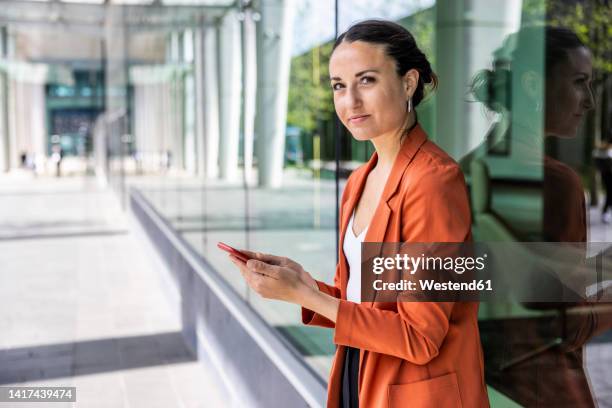 businesswoman holding smart phone leaning on glass wall - red clothes stock pictures, royalty-free photos & images