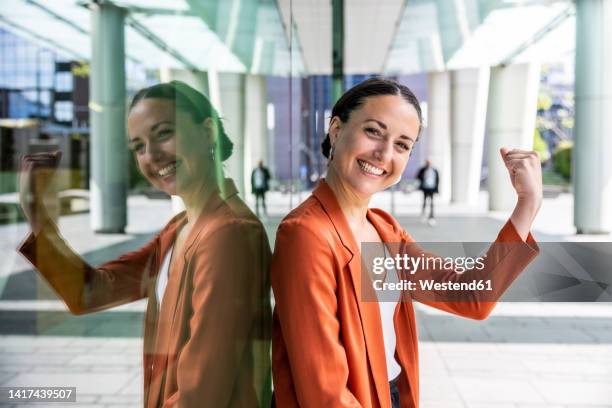 happy businesswoman showing muscle leaning on glass wall - flexing muscles stockfoto's en -beelden