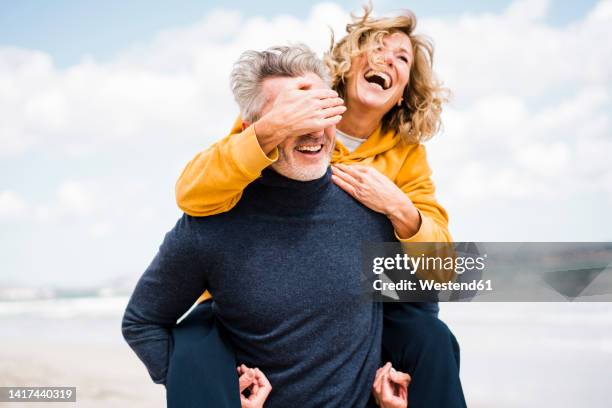 cheerful woman covering eyes of man with hand at beach - guess who stock pictures, royalty-free photos & images