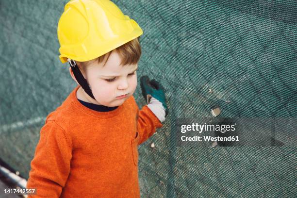 boy with hard hat standing by fence - boy in hard hat photos et images de collection