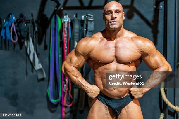 confident bodybuilder with arms akimbo flexing muscles in gym - body building stockfoto's en -beelden