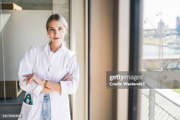 confident doctor with arms crossed leaning on wall - bata de laboratorio fotografías e imágenes de stock