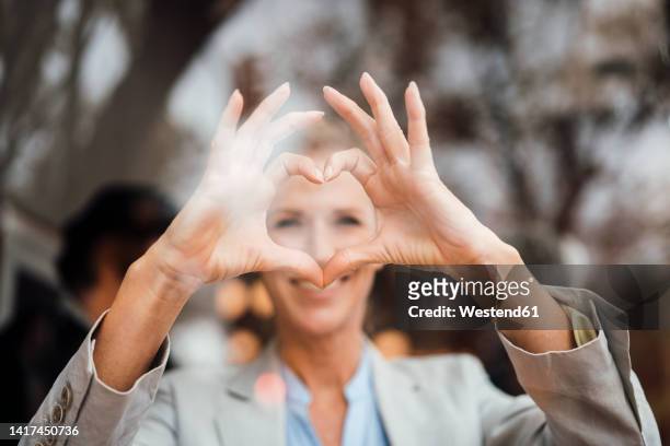 smiling businesswoman gesturing heart shape in cafe seen through glass - gestikulieren mit den händen stock-fotos und bilder