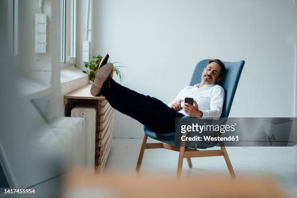 businessman holding smart phone sitting on armchair at office - smartphone office stock-fotos und bilder