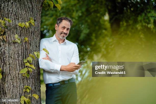 smiling mature businessman holding smart phone standing by tree - leaning tree stock pictures, royalty-free photos & images