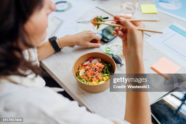 businesswoman eating healthy poke bowl with chopsticks in lunch break at office - pokes stock pictures, royalty-free photos & images