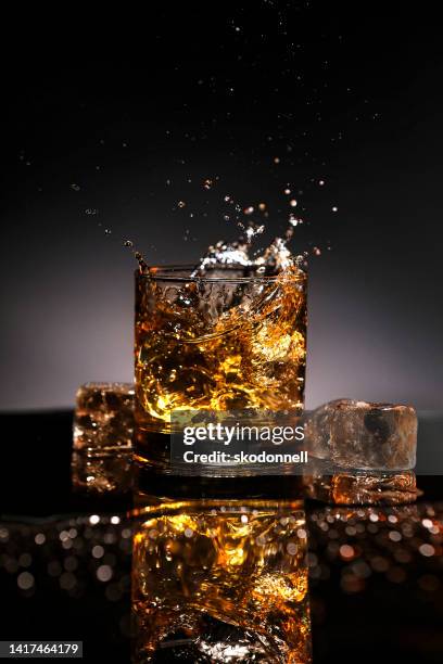 bourbon whiskey oder rum spritzt aus einem glas mit eis würfel auf schwarzer reflexion - whisky stock-fotos und bilder