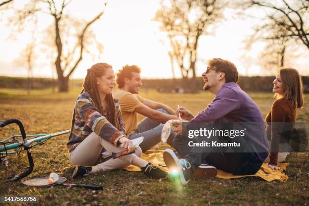 four young people sitting on a picnic blanket in the park, chatting and laughing - university student picnic stock pictures, royalty-free photos & images