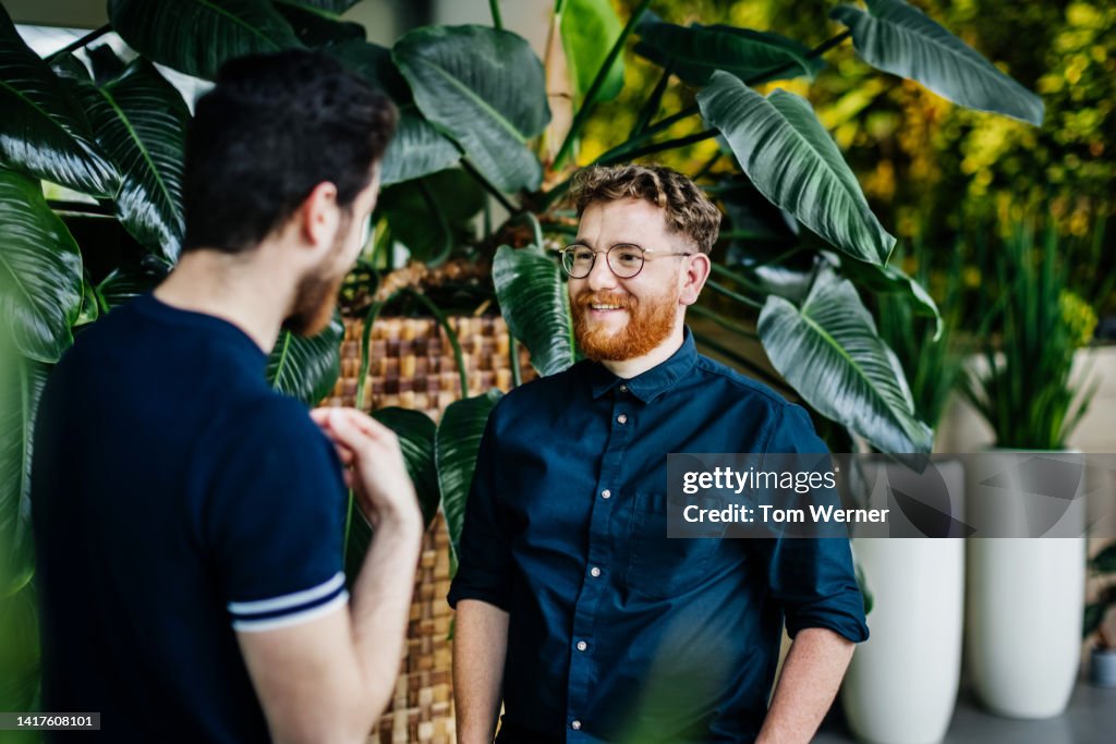 Two Colleagues Standing Amongst Potted Plants In Green Office Space, Talking