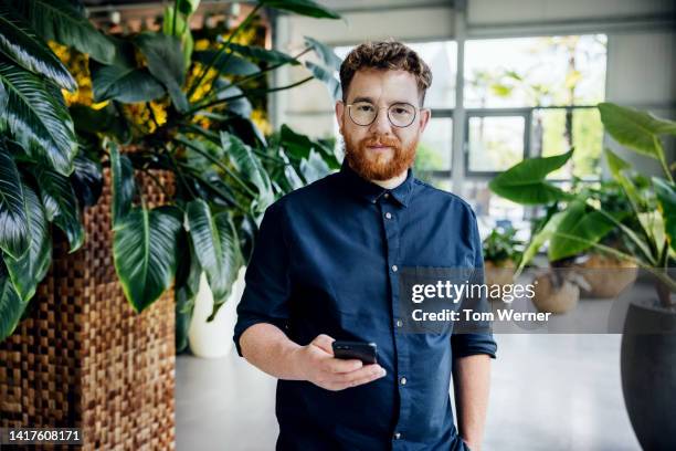 man with smartphone standing in large, open office space with lots of plants - business casual phone stock pictures, royalty-free photos & images