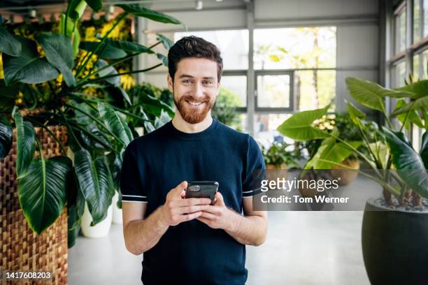 portrait of man smiling while using smartphone in office filled with plants - naturbursche stock-fotos und bilder