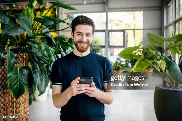 portrait of man smiling while using smartphone in office filled with plants - germany best pictures of the day stockfoto's en -beelden