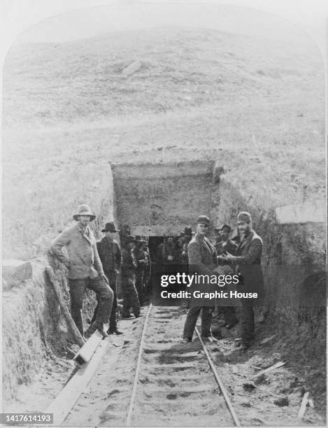 Coal miners stand around the entrance to a mine, around the tracks of a mine railway, a donkey pulling a cart emerging from the darkness of the...