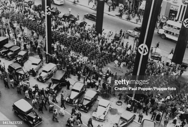Aerial view of spectators lining a street to watch a parade of German Army troops and brass band marching along an avenue lined with Nazi flags on...