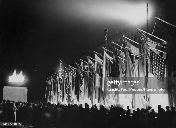 Crowds gather around a beacon containing the Olympic Flame beside the flags of participating nations flying from poles in front of the Altes Museum...