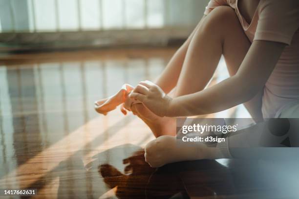 close up shot of little asian girl wearing socks and her ballet shoes by the window against sunlight while sitting on hardwood floor at home. getting ready for her ballet class. hobbies, interests and extracurricular activities for kids - dancing studio shot stock pictures, royalty-free photos & images