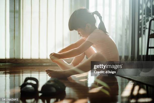 little asian girl wearing socks and her ballet shoes by the window against sunlight while sitting on hardwood floor at home. getting ready for her ballet class. hobbies, interests and extracurricular activities for kids - pointe stock pictures, royalty-free photos & images