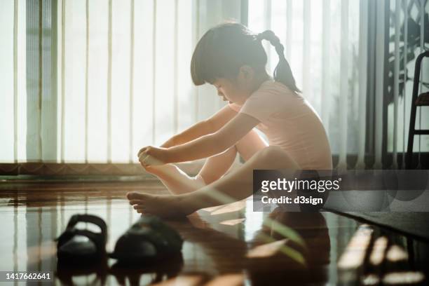 little asian girl wearing socks and her ballet shoes by the window against sunlight while sitting on hardwood floor at home. getting ready for her ballet class. hobbies, interests and extracurricular activities for kids - school gymnastics 個照片及圖片檔