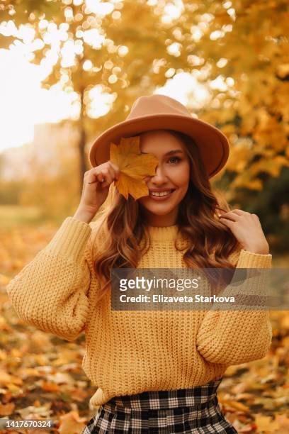 portrait of a beautiful young woman in a hat and a yellow sweater having fun and holding an autumn leaf enjoying solitude walking through the foliage in an autumn park outdoors in autumn. a pretty smiling girl in fashionable clothes is resting alone - single leaf stock-fotos und bilder