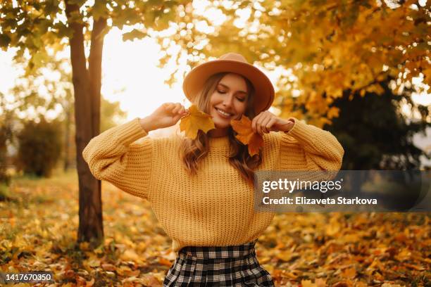 portrait of a beautiful young woman in a hat and a yellow sweater having fun and holding an autumn leaf enjoying solitude walking through the foliage in an autumn park outdoors in autumn. a pretty smiling girl in fashionable clothes is resting alone - september stock-fotos und bilder