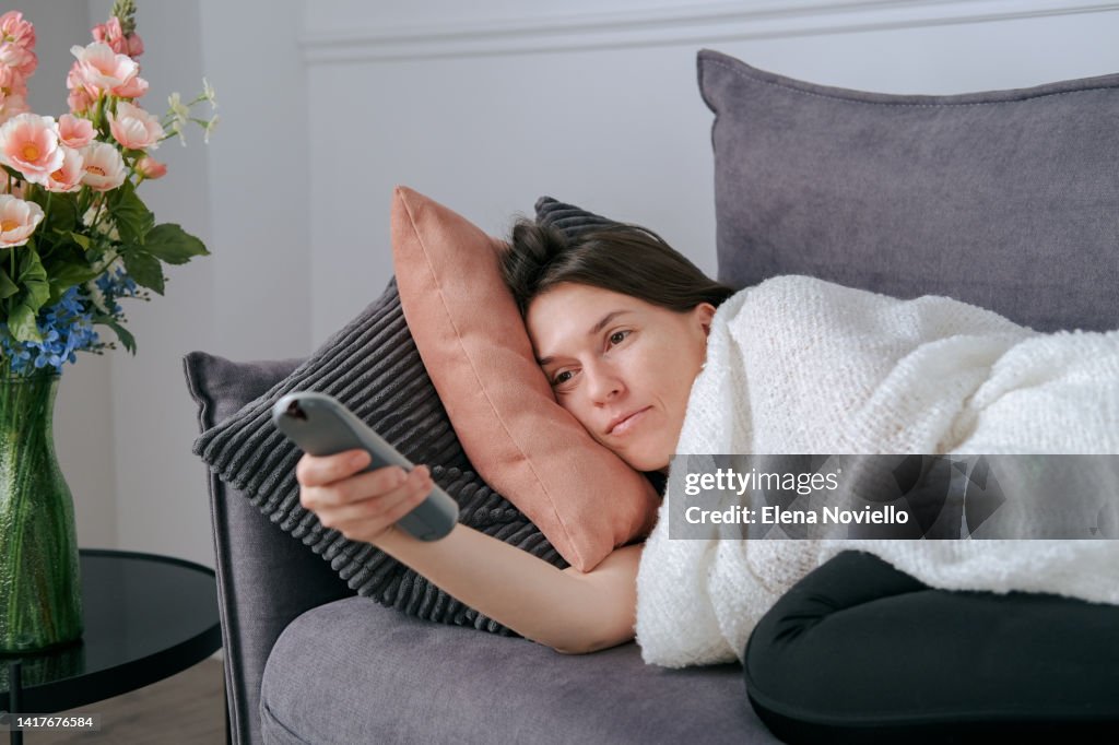 Cold at home young woman on the couch, covered with a warm blanket and switching TV channels, it's cold at home